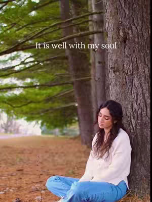A woman in blue jeans and a white sweater sits cross-legged, leaning against a large tree trunk in a forest. The background includes other trees and a soft, blurred green. Text overlayed on the top left says, 'It is well with my soul.'