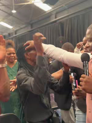 A blurry image of a group of people in what appears to be a church setting. A person, possibly a pastor, has his hand on the head of a person dressed in black.