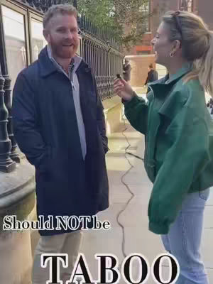 A woman in a green jacket interviews a man in a dark coat, positioned on a sidewalk or street with a metal gate and buildings in the background.