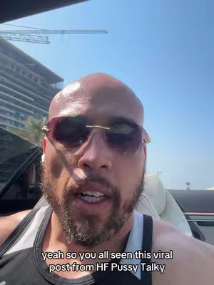 A selfie-style video shot from a low angle inside a vehicle. The main subject is a bald man wearing sunglasses and a tank top, with a visible beard and a serious expression. He is talking, and there's a textual overlay at the bottom of the screen. The background includes a bright blue sky, parts of a building, and a construction crane.