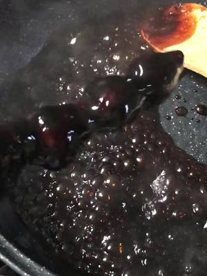 An overhead shot captures a pan filled with a dark, syrupy substance, likely a sugary glaze or sauce. Bubbles are visible, indicating heat and the ongoing cooking process. A wooden spoon rests within, with a small area of the dish in a more vivid reddish hue.