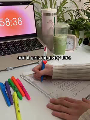 A well-lit workspace with a laptop showing a timer, a pen in hand writing in a notebook, with several colorful markers, a tumbler, and a cup of green drink. Plants add a touch of nature.