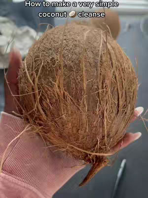 A close-up shot shows a large, shaggy coconut held in a person's hand. The coconut fills most of the frame, showcasing its rough, fibrous exterior. The hand holding the coconut is seen from the wrist up, with the person wearing a light pink-colored sleeve. The background is blurred, adding depth to the image.