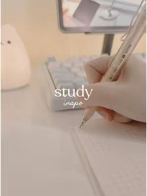 The image shows a desk setup with a hand holding a pen above a notebook. The color palette is soft and warm, mainly consisting of light beige and white, giving a cozy aesthetic. Elements include a computer monitor, a white keyboard, and a decorative object (possibly a lamp) on the left. The focus is on the act of writing, suggesting study or note-taking.