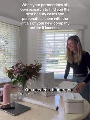 The image shows a domestic interior with a woman in the foreground smiling. She is standing near a table with a laptop, wrapping paper, and beauty cases. The background includes windows with blinds and a vase of flowers. Overall, the image has a well-lit, casual vibe.