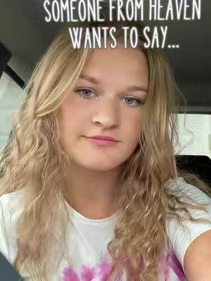 A head-and-shoulders shot of a young woman with long, wavy blonde hair. She has fair skin, blue eyes, and is wearing a white t-shirt. Above her, the text: 'SOMEONE FROM HEAVEN WANTS TO SAY...' is overlaid in a whimsical font, against the backdrop of a car interior.