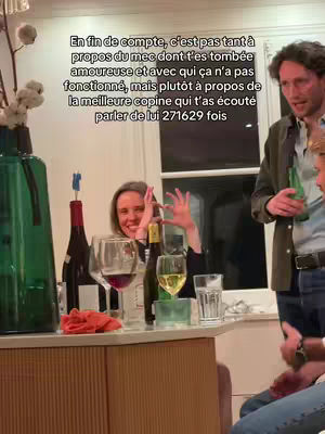 A candid scene depicting a group of people at a gathering, likely a house party or dinner. The main focus is on a woman at a table. She is smiling and holding up her fingers in a playful gesture. In front of her are wine glasses and bottles, adding to the relaxed, social atmosphere. Behind her are two men and the background contains a wall with a window.