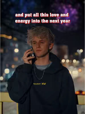 A young man with light blonde hair is on a phone call outdoors at what appears to be night. The background is softly blurred with city lights and a dark sky. The man is wearing a black hoodie. The text overlayed on the top reads, 'and put all this love and energy into the next year' in a red and white box.