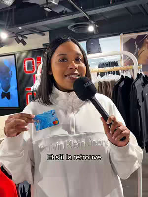 A woman stands indoors in a store holding a credit card and a microphone. She is wearing a white Calvin Klein Jeans jacket. Text is overlaid on the image.