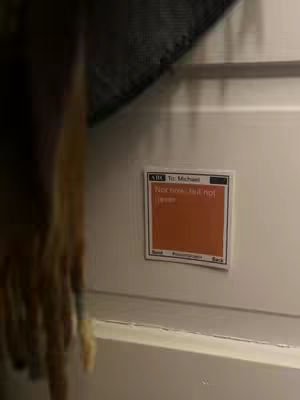 A close-up shot shows a square sticker attached to a white surface, likely a door. The sticker has a white border with text at the top, the center contains an orange fill. The foreground shows a blurry figure on the left. The background includes a dark, undefined area above.