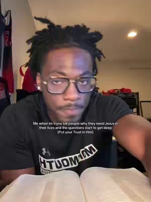 A close-up shot of a young Black man wearing glasses and a black t-shirt, sitting in front of a desk. A Bible is open in front of him. Text overlayed says, "Me when im tryna tell people why they need Jesus in their lives and the questions start to get deep (Put your Trust in Him)"