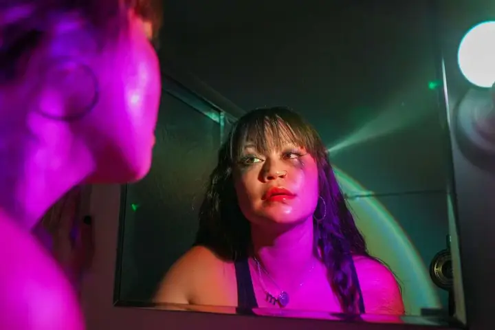 A person's face in a dimly lit bathroom, seen through a mirror. The face has makeup, including red lipstick and what looks like smeared eye makeup. Bright, colorful lighting casts purple and green hues on the scene, creating a dramatic effect. The person in the mirror is looking slightly away.