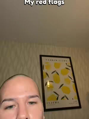 A close-up shot of a person's face, with text overlayed. The person has short hair and light skin. In the background, there's a framed picture with a yellow and white pattern, and a wall with a textured pattern.