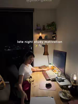 A woman is sitting at a desk studying at night. The desk is cluttered with a computer, keyboard, mouse, and other study materials. The background is dark, with a window looking out at a city skyline. The overall composition gives off a cozy and studious vibe.