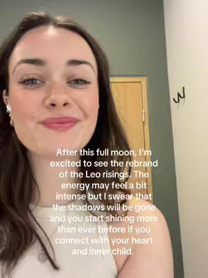 A selfie-style video with the creator's face filling most of the frame. The background includes a portion of a light-colored wall, a wooden door, and a decorative hook. The creator, a woman with brown hair, is smiling and looking towards the camera. Text overlayed on the image provides context related to a 'full moon' theme and astrology.