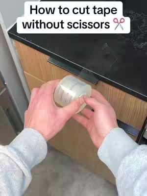 A first-person perspective shows a person holding a roll of clear packing tape. The person's grey sleeved arms and hands are in the center, with the tape roll held in the palm. The background shows a wooden cabinet with a black countertop. The top of the image has text overlay.