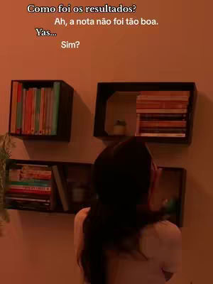 A woman is standing in front of a wall with three shelves holding books. The scene is dimly lit with a warm, orange hue.