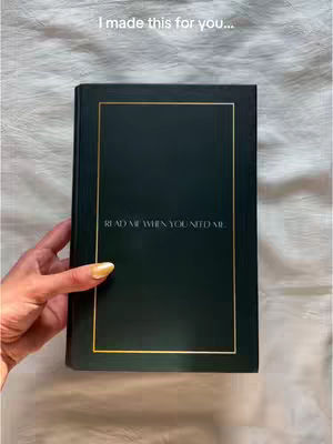 A hand holding a dark green journal or notebook against a textured white background. The book has a gold border, and the text 'READ ME WHEN YOU NEED ME' is visible on the front cover. Additional text 'I made this for you...' is at the top.