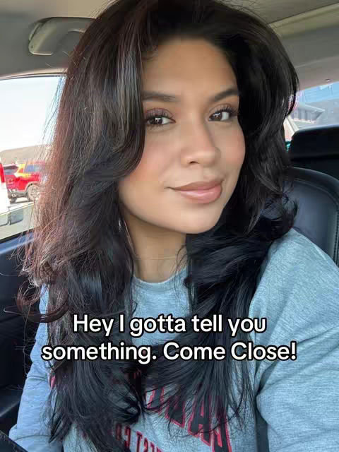 A woman with long dark hair is inside a car, with a friendly expression. The lighting is natural, and the focus is on her face. The background is blurred, with glimpses of a road and another vehicle.