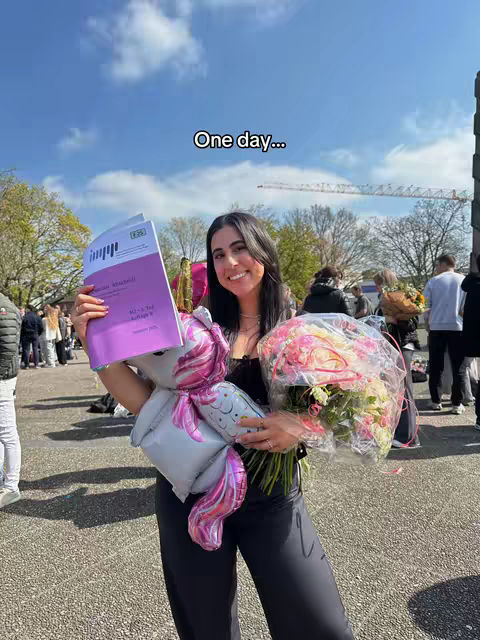 A young woman stands on a sunny day holding a book, a unicorn balloon, and a bouquet of flowers. She's smiling at the camera. Other people and a building are in the blurry background. The sky is blue with clouds.