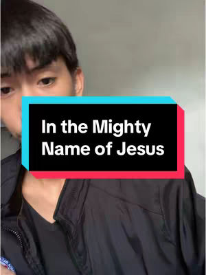 A person is visible from the chest up, with the frame centered on their face. The individual has short black hair and is wearing a black jacket. Overlaying the person is a rectangular box in a light color, with red accents on the top and bottom. Inside the box, the text 'In the Mighty Name of Jesus' is displayed in a bold, white font.