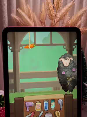 A colorful mobile game screenshot displayed on an iPad. The game scene shows a cartoon sheep with a black ruffled coat and a white face standing in front of a wooden fence. A carrot hangs from the top, and a shelf below displays styling tools.