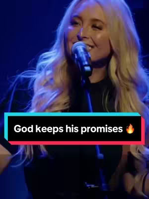 A woman with long blonde hair is singing into a microphone. She is wearing a black top and is positioned center frame. The background is dark, with blue and purple lighting. A text overlay at the bottom states \"God keeps his promises\" in white text over a black and red box, with a flame emoji.