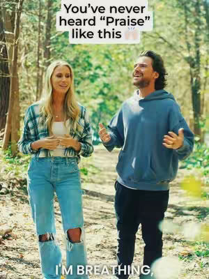 A medium shot captures a man and woman walking along a wooded path, sunlight filtering through the trees. The woman stands on the left, dressed in a plaid shirt and jeans with ripped knees. The man is on the right, wearing a blue hoodie and dark pants. Text overlay stating, 'You've never heard "Praise" like this' appears against a light-colored banner. The overall composition presents a casual, outdoor setting.