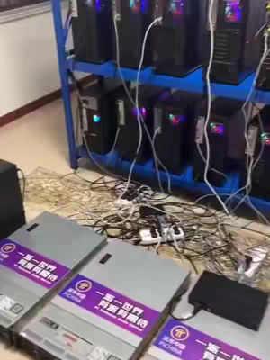 The image shows a room with multiple computer towers arranged on shelves and on the floor. Cables and power strips are visible, suggesting a setup for cryptocurrency mining or a similar technological application. The overall color scheme is a mix of black, blue, and the metallic tones of the computer hardware.
