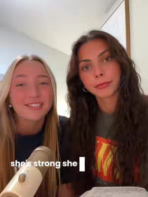 Two young women are positioned closely together, facing the camera. The woman on the left has long, light blonde hair and is smiling. The woman on the right has long dark hair and also smiles. A microphone is in the foreground. The background is a lightly colored wall and wooden trim with natural lighting, complemented by simple and neat features.