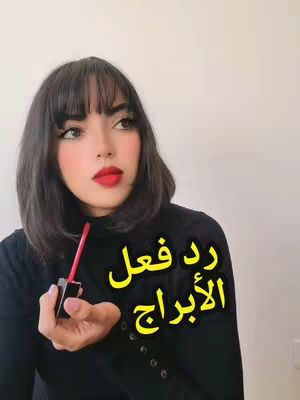 A woman with dark short hair and red lipstick holds a tube of lipstick in front of a white background. Text overlayed in yellow reads 'رد فعل الأبراج'.