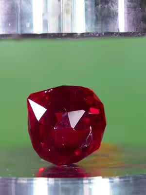 The image shows a large, faceted, ruby-red gemstone in the center, resting on a reflective surface. Above, a part of a metallic structure is visible, potentially a hydraulic press or similar machinery. The background is a solid green color. The composition is simple, emphasizing the gemstone with the suggestive implication of being crushed.