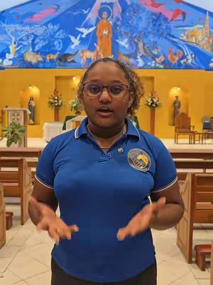A woman with glasses stands in front of a church interior. Behind her is a mural depicting religious figures with golden lights. The woman is speaking, with her hands up, as if presenting or explaining something.