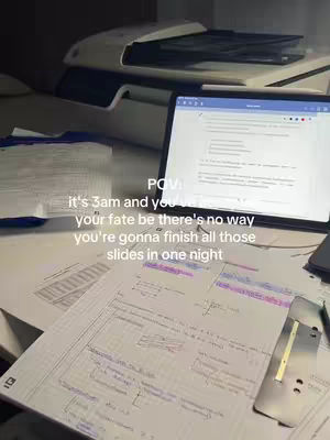 A dimly lit desk covered in study materials. A laptop screen displays text, and papers with handwritten notes are spread around. A printer sits in the background, and the overall scene suggests a late-night study session.