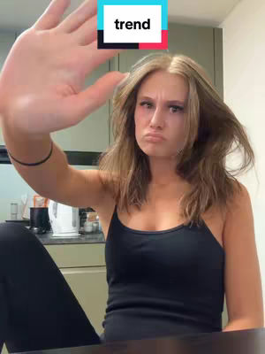 A young woman is shown in a casual setting, possibly her home. She holds her hand up towards the camera, creating a stop gesture with her palm facing forward. She wears a black tank top and has her hair down. A text overlay, labeled 'trend,' is placed in the upper center of the image, and she has a slightly dissatisfied expression.