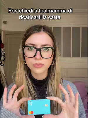A young woman with blonde hair and glasses holds up a turquoise credit card in front of her. She's in a well-lit room with a neutral background, suggesting a casual setting. Text overlayed at the top says, "Pov chiedi a tua mamma di ricaricarti la carta," indicating a video related to banking or finances.
