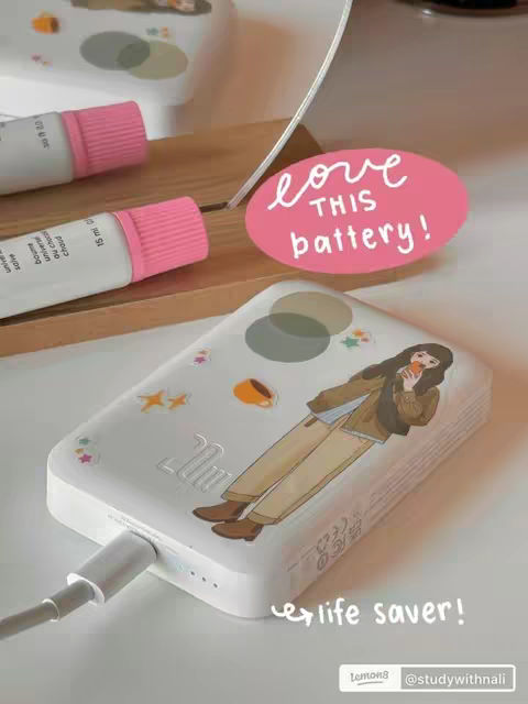 The image features various elements arranged on a light-colored surface, appearing to be a table or desk. In the foreground, a stylized power bank is prominently displayed. On the power bank is a cartoon image, featuring an illustration of a girl holding a container. Adjacent to the power bank are several small tubes with pink caps, arranged on a wooden surface. In the background, a pink oval sticker displays the text "love this battery!". The overall composition is clean and aesthetically pleasing, conveying a sense of both functionality and playfulness.