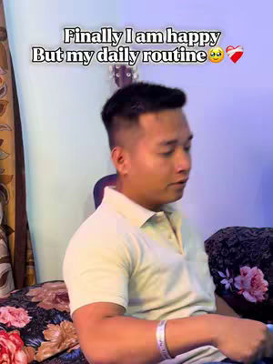 A medium shot features a young man sitting and looking to the side with a thoughtful expression. He is wearing a light-colored polo shirt and has short, dark hair. The background includes a blurred interior with a guitar visible and a patterned floral sheet. A text overlay appears at the top, and the overall composition is somewhat muted, implying a moment of quiet reflection.