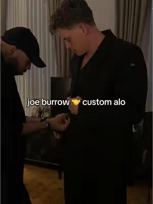 The cover image captures Joe Burrow in a dark, long coat being attended to by another person, possibly adjusting his clothing. The setting appears to be a well-lit interior, with a dark background and visible light fixtures, the primary focus is on the two figures as they're framed from the waist up . The text overlay indicates the outfit is custom Alo.