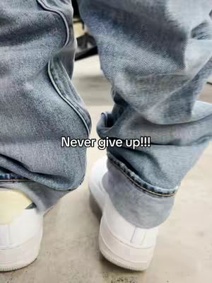 The image shows a person's legs from the knees down, wearing light blue jeans and white sneakers. The jeans are slightly baggy, and the bottom of the legs are scrunched up around the ankles. The sneakers are plain white, and the background is a light gray surface. Text overlay: Never give up!!!