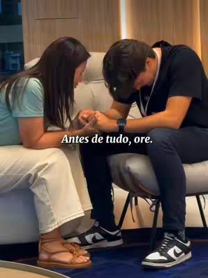 Two individuals, a woman and a man, are seated close together on armchairs, holding hands in prayer. The woman is to the left, in a light blue shirt and white pants, with medium length dark hair. The man, to the right, is wearing dark pants and a black top, with short, dark hair. The text overlay reads, 'Antes de tudo, ore.'
