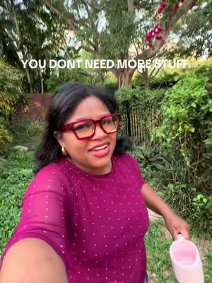 A woman with dark hair and red-framed glasses stands outdoors. The background shows lush greenery with a large tree and some flowering plants, and the woman holds a pink container in her right hand, while she smiles at the camera, conveying a positive and engaging message.