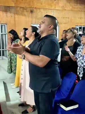 A medium shot captures a group of people, presumably in a church setting, with the main focus on a man standing center-right. He is the most prominent figure, with his hands raised in a gesture of worship or praise, mouth open as if singing. The background is softly blurred, suggesting a spacious room with a wooden clad background.