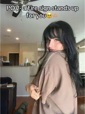 A young woman with long black hair and bangs is shown in front of a home interior; she is facing slightly away from the camera, with her arms crossed. She is wearing a loose beige top and turning her head back to the camera with a slight smile. Behind her, interior details such as a kitchen counter, a ceiling fan, and a wooden floor are visible. The background is well-lit, suggesting a bright daytime setting.