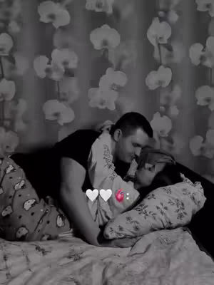 A black and white image of a couple in bed, hugging and kissing. The background features a floral pattern.