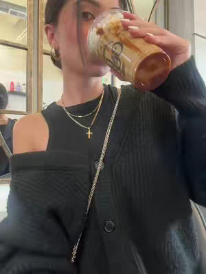 A woman is shown in a coffee shop, holding a coffee cup to her mouth. She is casually dressed, with layered gold necklaces and a cross pendant, and has her hair pulled back. The background features a portion of a glass display case and wall.
