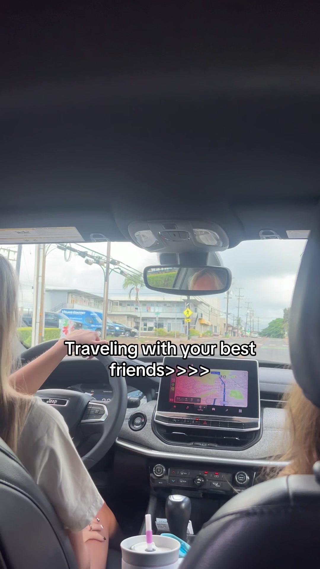 The image is shot from inside a car, likely a Jeep, with two blonde-haired women visible. One is driving, her arm resting on the steering wheel. Text overlayed on the image reads 'Traveling with your best friends>>>', likely implying a travel vlog or story about friendship. The car interior is visible, along with an outdoor setting with road and buildings.