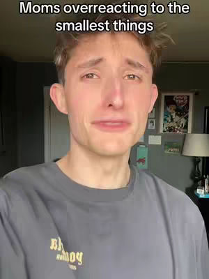 A young man with light brown hair is crying, appearing distressed. Text overlayed above the figure reads 'Moms overreacting to the smallest things'. He's wearing a gray t-shirt with a graphic on it. The background includes a glimpse of a room with a framed picture hanging on the wall. The lighting appears to be natural.