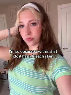 A selfie-style shot featuring a young woman. She has long, wavy hair with blonde highlights near her face, and is wearing a white headband. She's wearing a light green striped shirt. The background is somewhat blurred, showing what appears to be a room with doors and a piece of wall decor.