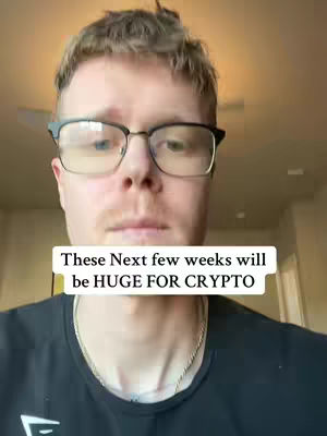 A man with short brown hair and glasses is centered in the frame. Above his dark t-shirt, a white rectangle with black text appears: 'Next few weeks will be HUGE FOR CRYPTO'. The background is simple, with neutral tones and a hint of a ceiling fan. The shot is a face close-up, focusing on his expression and messaging.